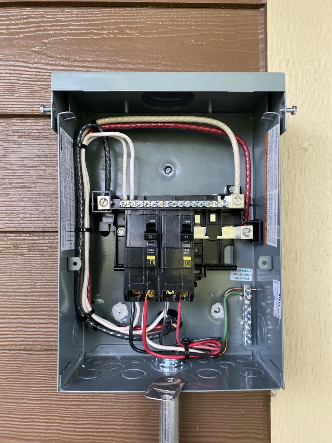 A close-up view of an open electrical circuit breaker box mounted on a wall, showing various wiring connections and labeled breakers.