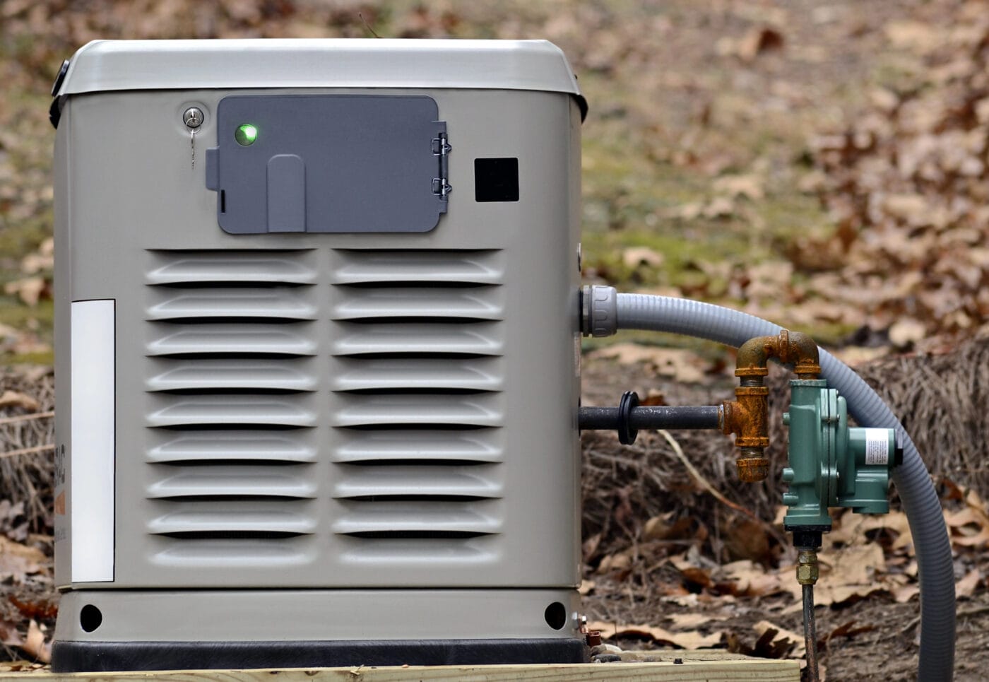 Home standby generator with a gray exterior, connected to a gas meter and pipes, surrounded by dried leaves on the ground.