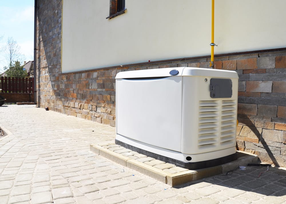 A large, white outdoor generator is placed next to the stone wall of a building on a paved area.