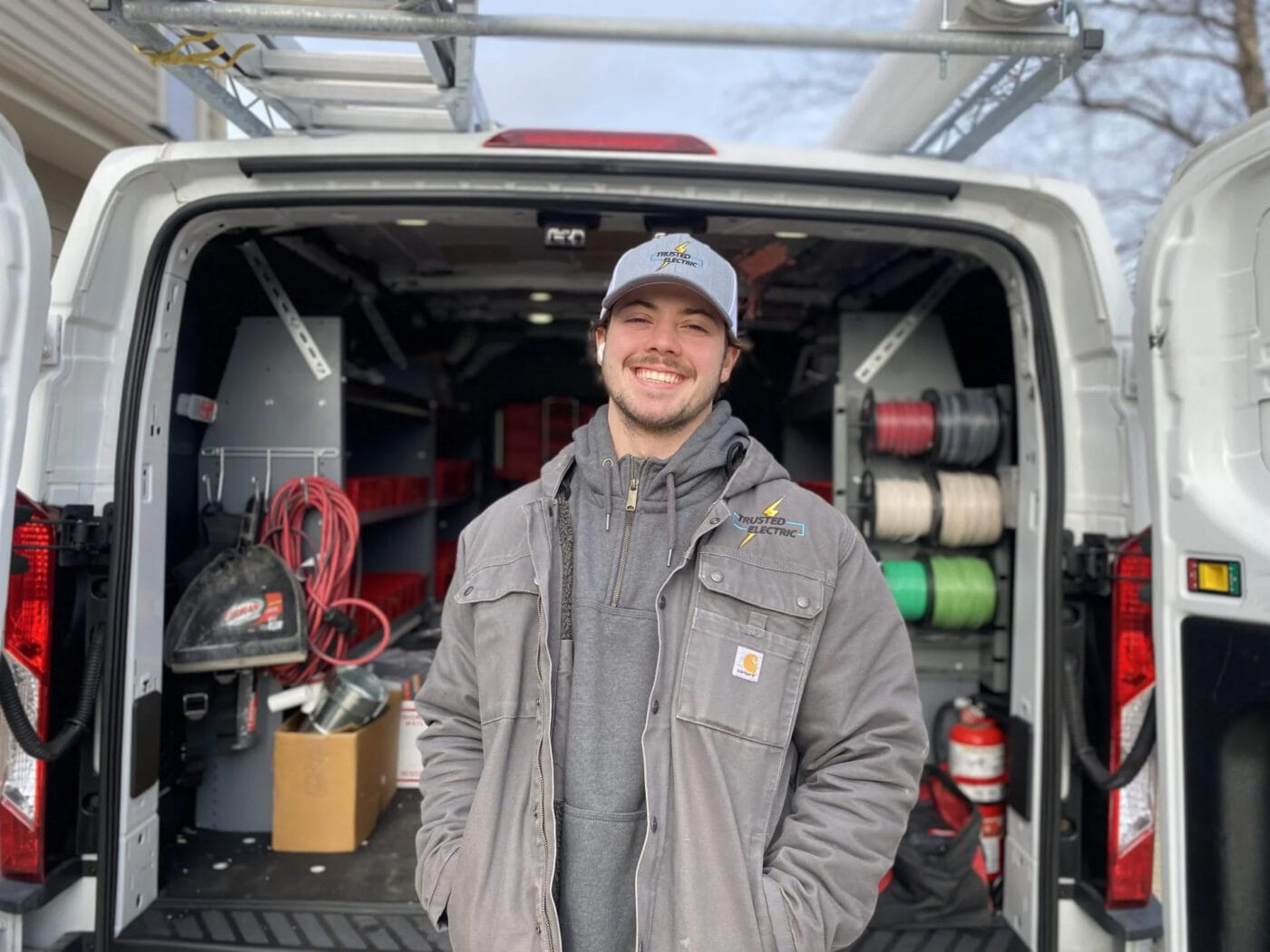 Owner of trusted electric stands smiling in front of an open work van filled with tools and equipment.