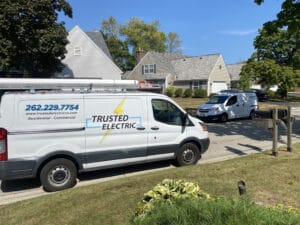 Two white electrical service vans are parked on a suburban street in front of a house with a grey roof.