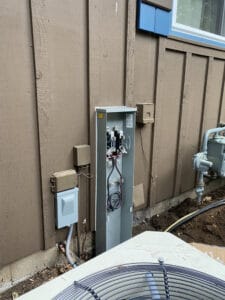 Outdoor utility setup with an open electrical box showing wires and components, next to other utility boxes on a wooden wall near a window and an AC unit.
