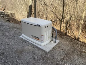 A Generac generator is installed on a concrete slab in a wooded area, surrounded by trees without leaves.