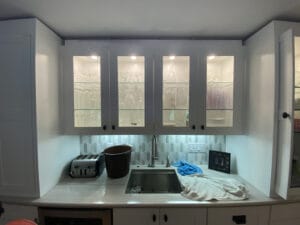 Kitchen sink with overhead glass cabinets displaying dishes, a black pail, a cloth, a toaster, and a decorative frame on a countertop.