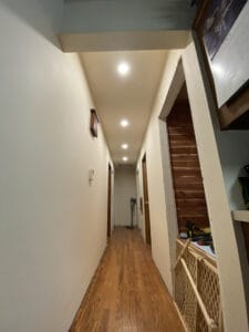 A narrow hallway with wooden flooring, white walls, and ceiling lights. Doors on both sides lead to different rooms. A baby gate and tools are seen on the right.