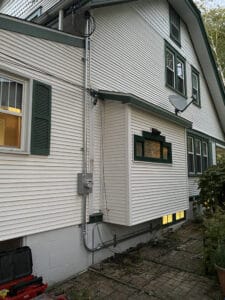 Side view of a white house with green trim, featuring several windows, a satellite dish, and electrical wiring on the exterior wall.