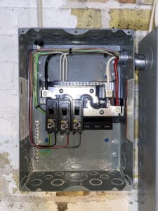 Electrical panel with exposed wiring, featuring three circuit breakers and multiple colored wires on a brick wall.