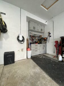 A garage corner with a metal storage cabinet, various tools hanging above, a black mat on the floor, and a sealed box in the foreground.