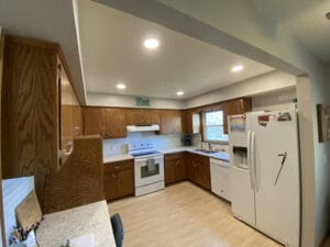 A kitchen with wooden cabinets, a white stove and refrigerator, and light wood flooring. The counters have various small items, and there is a window above the sink.