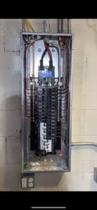 Electrical panel with exposed wiring and breakers against a white brick wall.