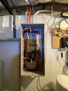 An open electrical panel in a basement shows various colored wires and components. The ceiling has visible insulation and pipes.
