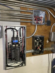 Electrical panel with wires and circuit breakers mounted on a wall, alongside two smaller open electrical boxes showing additional wiring.