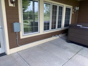 Outdoor patio with brown siding, three windows, an electrical box, and a covered hot tub on a concrete surface. Trees and grass are visible through the windows.