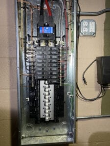 An open electrical panel with visible wiring, circuit breakers, and a few closed switches. Nearby, a dual electrical outlet is mounted on the wall.