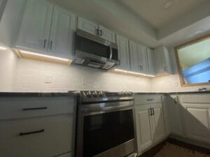 Modern kitchen with white cabinets, under-cabinet lighting, a stainless steel oven, and a built-in microwave. Subtle lighting highlights the tiled backsplash.