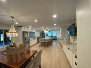 A spacious kitchen with a long, central dining table, white cabinets, stainless steel appliances, and wooden flooring. A chandelier hangs above a side table with a cardboard box.