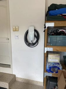 An electric vehicle charging station with a coiled cable mounted on a white wall beside a wooden shelf in a garage.