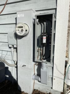 An outdoor electrical meter and breaker box mounted on a house exterior with visible wiring connections.