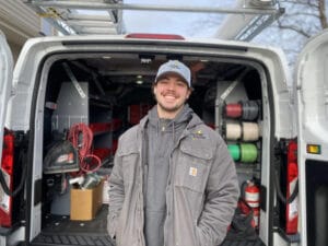 A person in a gray jacket and cap smiles while standing in front of a van filled with equipment and supplies.