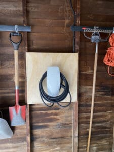Electric vehicle charger mounted on a wooden board in a garage, flanked by a shovel on the left and a broom and coiled orange extension cord on the right.