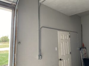 A gray wall with electrical conduits leading to an outlet near a closed door. A window on the left and a sign with partial text beside the door.