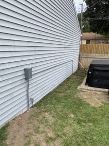 A side view of a house with gray siding, showing an electrical box and wires. There's grass and a black rectangular object in the foreground.